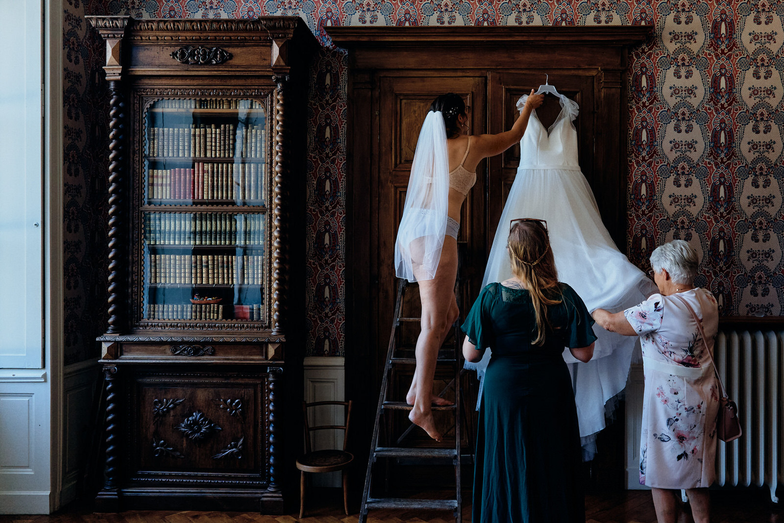 Mariée attrapant sa robe perchée sur un escabeau avec l'aide de sa grand-mère et d'une amie