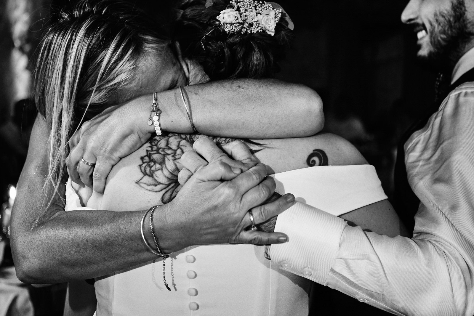 Photo de mariage en noir et blanc d'un calin plein d'émotions entre la mariée et une invitée, le marié la réconfortant d'une main dans son dos