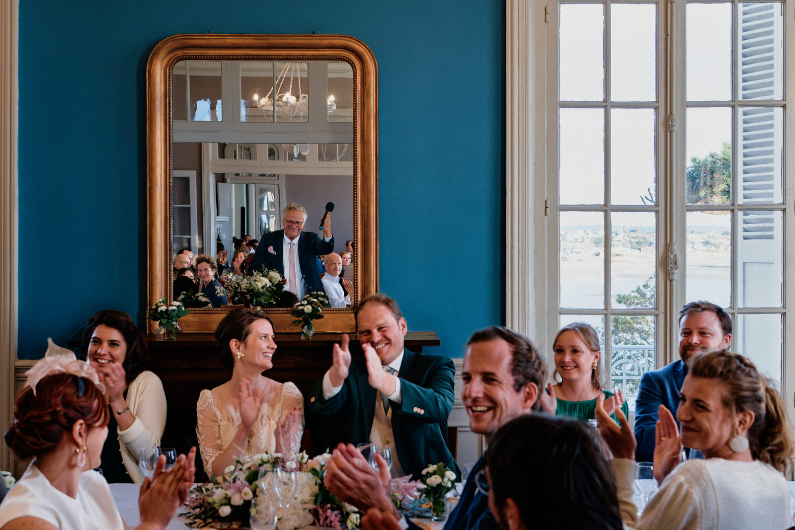 Fin d'un discours pendant une soirée de mariage. On voit les mariés et leurs invités, mais aussi la personne qui a fait le discours dans un miroir