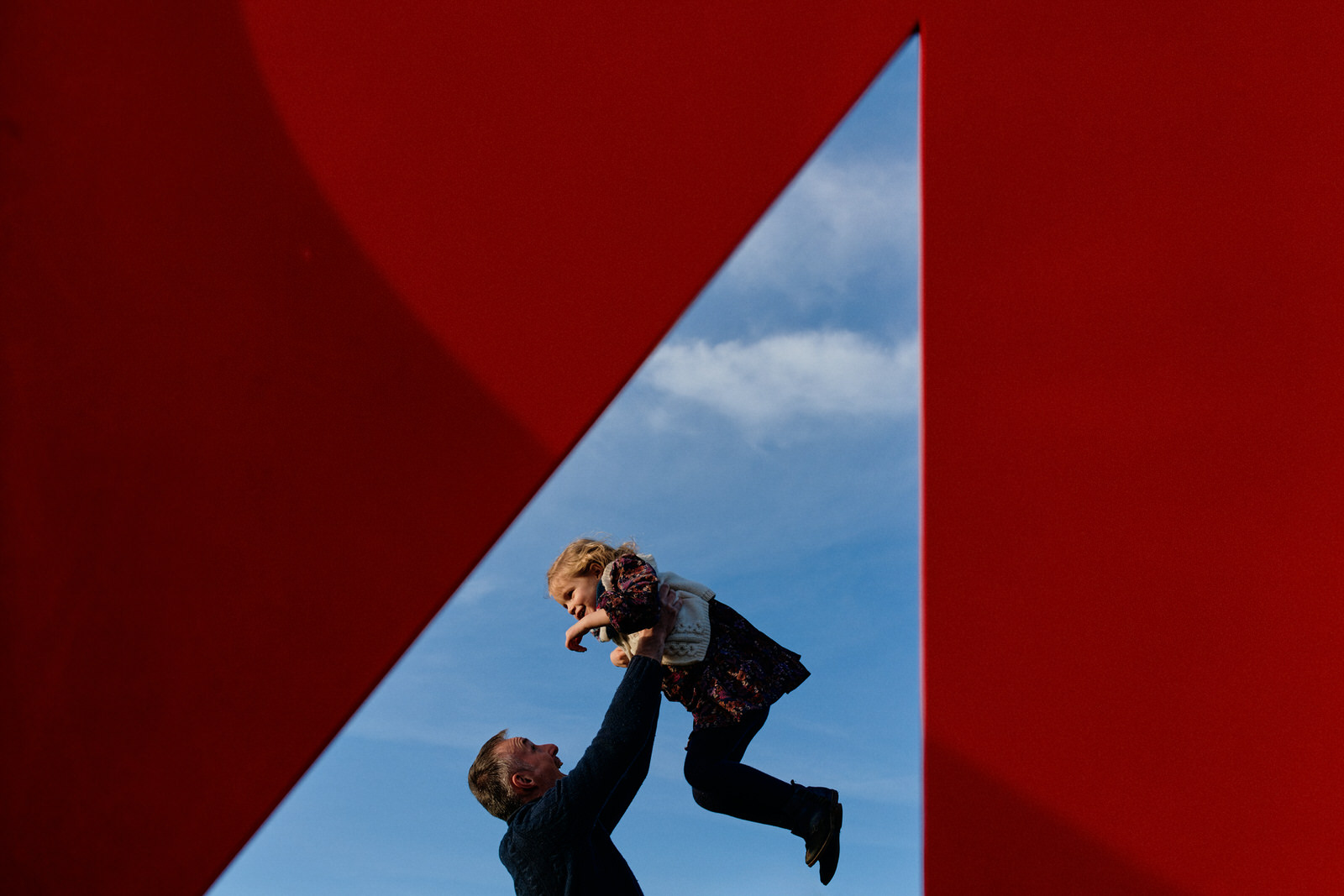 Photo d'un papa faisant voler sa fille en l'air, prise à travers du mobilier urbain donnant un aspect graphique et minimaliste à l'image