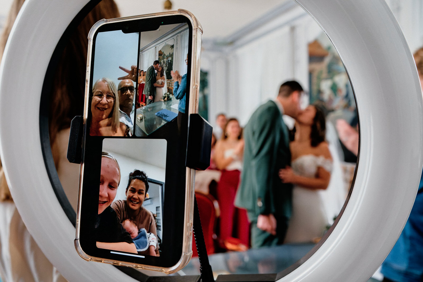 Couple de mariés s'embrassant à la mairie, la photo est prise à travers le pied tenant un téléphone sur lequel la famille est en visio, la mise au point est sur le téléphone mais on devine le bisou des mariés légèrement flou à l'arrière-plan