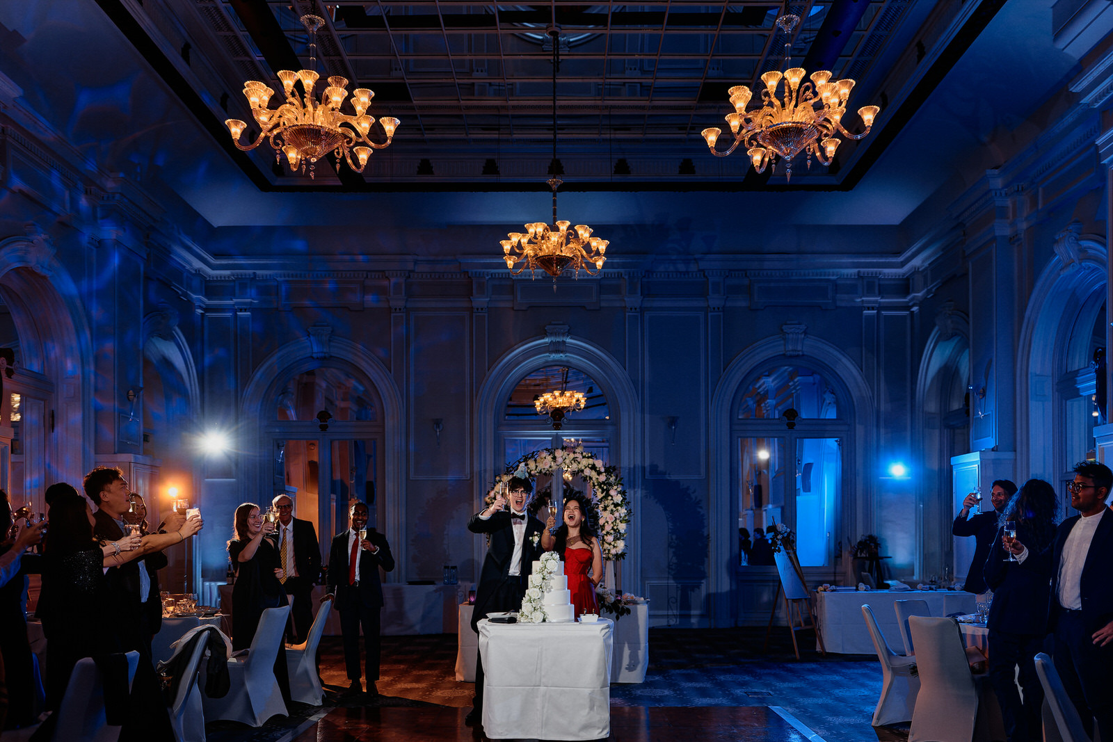 Photo d'un couple de mariés devant leur gâteau de mariage pendant la soirée, dans la belle salle d'un hotel, trinquant avec leurs invités