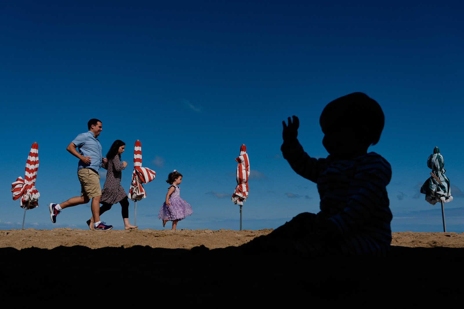 Séance famille sur une plage. Au premier plan la silhouette du bébé, et à l'arrière plan la grande soeur et les parents qui courent entre des parasols colorés