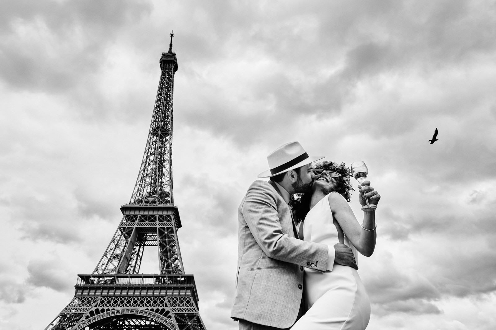 photo de couple de marié devant la tour eiffel, ils s'embrassent, la mariée tient un verre de champagne, un oiseau passe dans le ciel