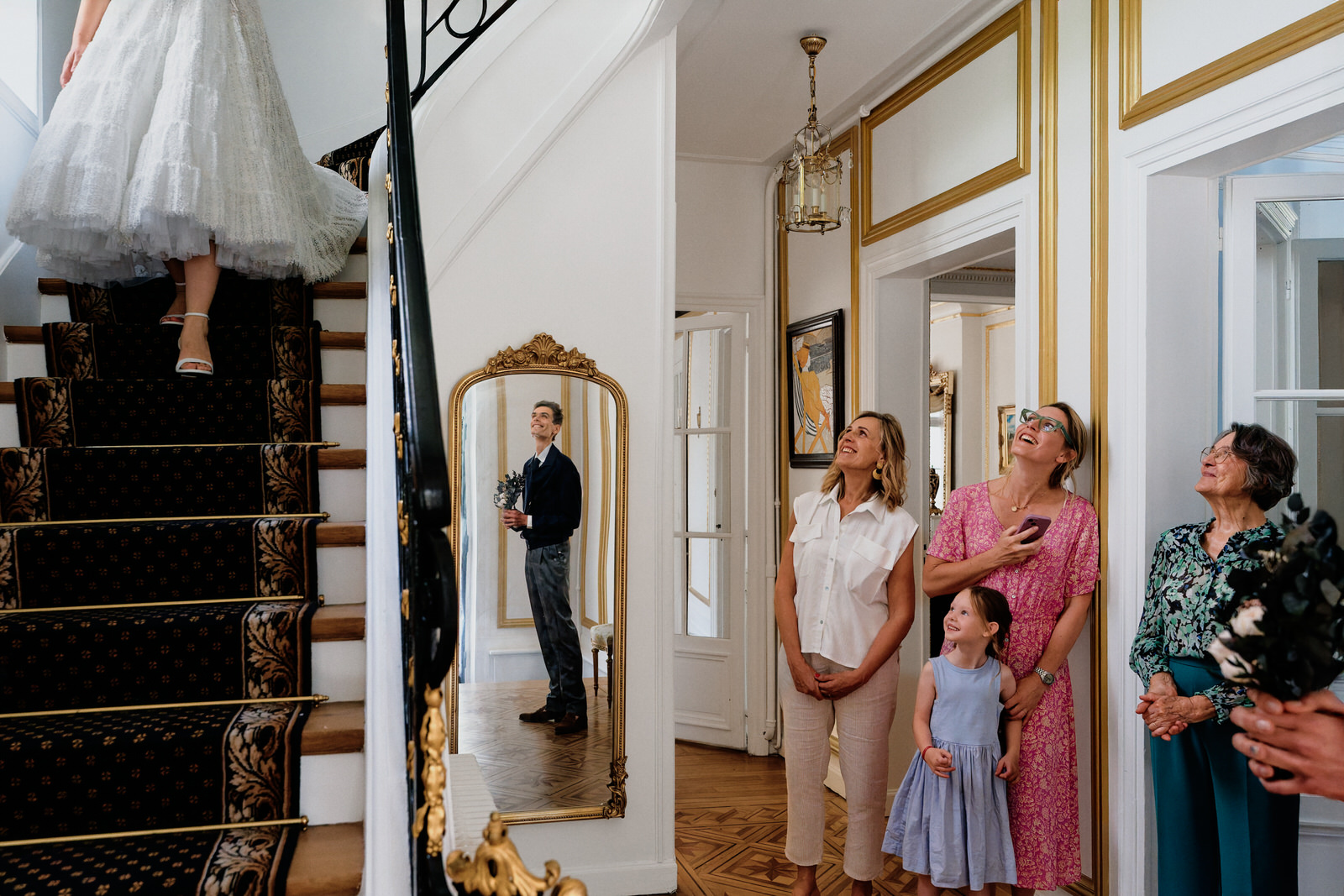 la mariée descend les escaliers, en bas on voit le marié en reflet dans un miroir et toute la famille du marié qui la regarde en souriant